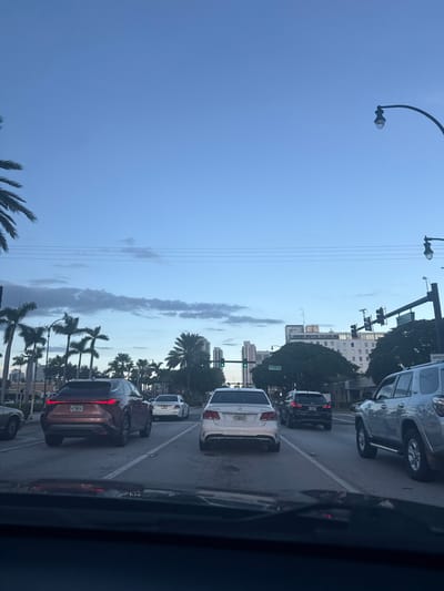 The image depicts a multi-lane street scene in Hallandale Beach, United States, viewed from the interior of a stationary vehicle during twilight conditions. The sky above is a clear, light blue with scattered, wispy grey clouds, transitioning to a deeper blue overhead. Multiple utility lines traverse the upper portion of the frame.
The roadway, likely asphalt, shows distinct white lane markings, including solid and dashed lines. Three primary vehicles are positioned directly ahead of the observer:
1. In the leftmost lane, a dark copper or bronze Lexus RX SUV is stopped. Its taillights are not illuminated.
2. In the center lane, a silver or white Mercedes-Benz E-Class sedan is stopped. Its taillights are not illuminated.
3. In the rightmost lane, a silver Toyota 4Runner SUV is stopped.
Further down the road, additional vehicles are visible in all lanes, indicating traffic congestion or a stop at a signal. A white sedan is visible behind the Lexus, and a dark SUV is behind the Mercedes.
The environment features numerous palm trees lining both sides of the street, consistent with a Florida landscape. In the background, a cluster of multi-story buildings forms a city skyline, including a prominent light-colored building with multiple windows. Streetlights with curved arms are present on poles along the road. Traffic light poles with multiple signal heads are visible, with a green light illuminated on a signal further down the road on the right side. A partial "SPEED" sign is also visible on the right. No individuals are discernible within the frame.