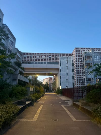 The image captures an urban architectural landscape at twilight, presenting a view dominated by large, geometrically structured buildings. The sky above is a clear, deep blue, indicating either dawn or dusk, with artificial lights already illuminating parts of the scene.
On the left and center-left, several modernist buildings rise prominently. The most striking structure features a facade composed of a repeating grid pattern of dark, recessed squares and lighter, flat rectangular sections, characteristic of Postmodern architecture, particularly reminiscent of Ricardo Bofill's work at Espaces d'Abraxas in Noisy-le-Grand, France. Windows are integrated into this complex grid. Another building, partially visible towards the top, also displays intricate concrete forms and window arrangements.
An elevated pedestrian walkway or bridge connects two sections of the buildings in the mid-ground, creating an archway or tunnel beneath it. Beyond this passage, additional buildings and dense foliage are visible, receding into the distance.
To the right, a paved path or road stretches away from the viewer, marked with distinctive light-colored lines forming rectangular shapes, possibly indicating parking or designated pedestrian zones. Along the edge of this path, a low wall or raised planter supports a vibrant array of green bushes and trees. Streetlights emit warm, yellow light, casting a glow on the path and the lower sections of the buildings. The scene appears devoid of people, conveying a sense of quiet grandeur. No visible text is present in the image.