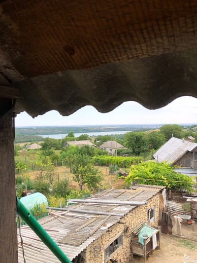The image depicts a view from a vantage point under a corrugated roof, looking out over a rural landscape. In the foreground, there is a weathered wooden beam and the undulating edge of a corrugated roof. Beyond this, a village is nestled amongst lush green trees. Several houses with grey or metallic roofs are visible, some with stone or light-colored walls. A body of water, likely a river or lake, stretches across the middle ground, with rolling hills covered in vegetation in the distance. The overall impression is one of a peaceful, somewhat rustic, rural settlement. The location is identified as the city of Calfa, Moldova.