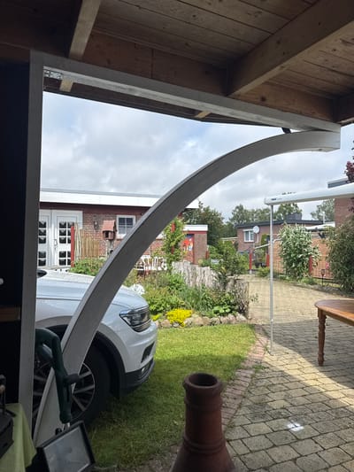 The image presents an outdoor scene captured from beneath a wooden overhead structure, likely a carport or patio cover, in Deinste, Germany. The structure's ceiling is composed of unpainted wooden planks and beams. A light grey painted wooden beam runs horizontally across the top left, with a metal hinge bracket visible attaching it to a vertical post on the far left, which is painted dark grey.

A prominent, large, light grey, curved wooden arch extends from the left side of the frame, arching towards the right and upward, connecting to the horizontal beam of the overhead structure. This arch forms a visual barrier and frames the view beyond.

Partially visible beneath the arch and to its left is the front passenger side of a white SUV, possibly a Volkswagen Tiguan, parked on a grassy area. Rain droplets are visible on the vehicle's surface. Underneath the covered structure, near the car's wheel, a green power tool with a handle (possibly a jigsaw or similar saw) is partially visible, leaning against a surface. A black floodlight sits on a lower surface in the bottom left foreground. A reddish-brown terracotta pot or chimney piece is visible in the bottom center.

Beyond the covered area, an outdoor garden environment is depicted under an overcast sky. A paved path made of grey interlocking bricks extends from the mid-ground towards the background, bordered by a strip of grass and various plants and small shrubs. To the right of the path, a light brown wooden outdoor table with a single visible ornate leg stands on the paving.

On the right side of the image, a white extendable awning or canopy, marked with "THULE Omnistor," is visible, supported by a metal pole extending to the paved ground. Further in the background, several houses with red brick exteriors and white window frames are discernible. One house in the mid-left features double white doors with multiple small glass panes. A reddish-brown wooden bird feeder on a post is visible in the garden area in front of this house. Various garden beds with green foliage and some yellow flowers are present. Trees are visible in the distant background.