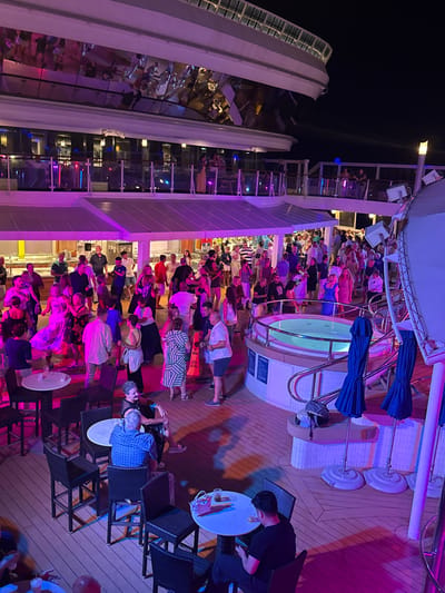 An aerial view captures a nighttime outdoor party on a cruise ship deck. A large crowd of people is engaged in various activities, including dancing, socializing, and sitting at tables. The deck is illuminated by vibrant pink and purple lights, creating a festive atmosphere. A circular hot tub is visible on the right side of the image, surrounded by railings. Several tables and chairs are scattered across the deck, some occupied by patrons. Blue umbrellas are positioned near the hot tub. The upper levels of the cruise ship, with reflective surfaces showing distorted images of people, are visible in the background. The scene appears to be a lively social gathering on a ship at sea, not in a city.