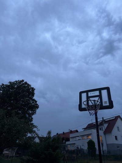 A basketball hoop with a clear backboard and red, white, and blue net stands in the foreground, positioned to the right of the frame. Behind the hoop, a residential area is visible. Several houses with red-tiled roofs and white walls are clustered together, some with lights on inside. A large tree with dark green foliage occupies the left side of the image, obscuring part of the sky and houses. The sky is overcast with heavy, dark grey clouds, suggesting an impending storm or the end of daylight. The scene appears to be a backyard or residential area in Schömberg, Germany, given the architectural style and presence of residential housing. There are no people visible in the image, and no actions or interactions are taking place.