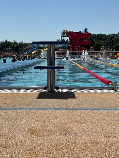 This is a photograph taken at an outdoor swimming pool, likely part of a water park. The scene is a wide shot of a swimming pool with lane markers, a diving block in the foreground, and people swimming and spectating. In the background, there are red water slides and a line of trees against a clear blue sky, suggesting a sunny day. The poolside is paved with a textured, light brown surface, bordered by a thin blue line. The water in the pool is clear and blue, reflecting the bright sunlight.
The focus of the image is on the pool area. In the foreground, a metal diving block is prominently featured, with its sleek, modern design. The pool itself is divided into lanes by red and yellow buoys. Numerous people are visible in and around the pool, some swimming in the lanes, others standing on the edge or by the slides. The water slides in the background are a vibrant red, indicating a recreational facility. The overall atmosphere appears to be one of leisure and summer fun.
The time of day is likely midday or early afternoon, given the bright sunlight and the activity at the pool. The weather is fair and sunny, conducive to outdoor water activities. There are no visible text elements in the image. The location context is Oława, Poland, implying the setting is a public pool or water park in that region. The presence of water slides and a crowd suggests it is a popular spot during warm weather.