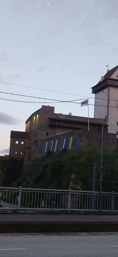 The image captures a view of Ivangorod Fortress in Russia, situated alongside a street and bridge railing. The fortress is constructed of dark stone, with a tall, imposing structure punctuated by windows. Flags are displayed along the fortress wall, including what appears to be Ukrainian flags and EU flags. Overhead, power lines stretch across the sky, which is light, suggesting twilight or early evening. A building stands adjacent to the fortress, and greenery is visible around the base of the structure. The road next to the railing and the overcast sky indicate a relatively cool or temperate weather condition.