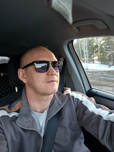 A man with a shaved head and wearing sunglasses is sitting in a car, presumably in the passenger seat. He is wearing a grey jacket with a white stripe on the sleeve and a seatbelt. The car appears to be in motion, and the scenery visible through the window includes trees and possibly snow or ice. According to the metadata, the location is Дулесово, Russia.