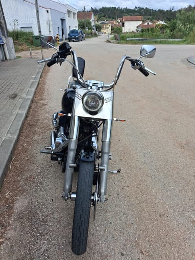 The image shows a motorcycle parked on an asphalt road in what appears to be a residential area of Soure, Portugal. The motorcycle is facing the camera, giving a clear view of its front end, including the headlight, handlebars, and front tire. The surrounding environment includes houses, greenery, and a curb along the edge of the road. The sky is overcast.