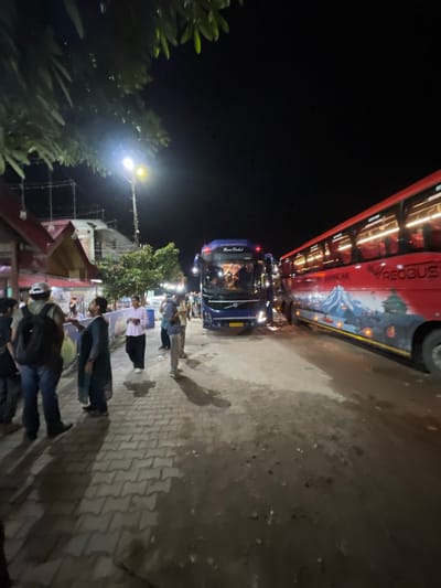 A night scene in Katauhar Kalan, India, captures a street with two parked buses and several individuals. On the left, a blue Volvo bus, identified by the text "Rani Dahal" on its front, is partially visible. To its right, a red bus with "PREOBUS" and mountain graphics is parked. The ground is a mixture of paved bricks and wet, muddy patches, reflecting the streetlights.

Several people are gathered in the area. On the left, near the blue bus, a person wearing a white cap and a backpack stands facing away from the camera. To their right, a woman in a dark outfit gestures with her hands. Further right, a person in a pink shirt and black pants walks across the scene. Another individual in a light-colored jacket is on their phone. More people are visible in the background, near the buildings.

The environment suggests a temporary stop or waiting area, possibly a bus station or a roadside gathering point. Overhead, trees with dark green leaves are silhouetted against the night sky, and a street lamp illuminates part of the scene. In the background, there are buildings with visible windows and a roof structure, along with scaffolding, indicating ongoing construction or renovation. The overall atmosphere is dimly lit, with the primary light sources being the buses' headlights and the street lamp.