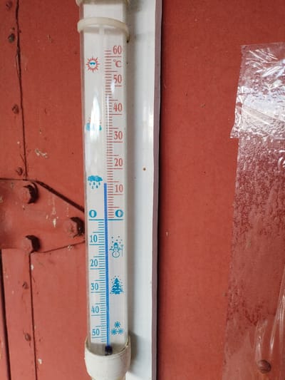 The image shows a thermometer mounted on a weathered red metal surface. The thermometer is a glass tube with markings in Celsius, ranging from -10 to 60 degrees. The red liquid level inside the thermometer is just above the 0 degree mark, indicating a temperature slightly above freezing. There are icons on the thermometer representing weather conditions: a sun with sunglasses at 60°C, raindrops at 10°C, and a snowman at -10°C, a Christmas tree at -30°C, and snowflakes at -50°C. The red surface appears to be part of an external structure, possibly a shed or a building's exterior wall. There is also a piece of clear plastic sheeting attached to the right side of the frame. The overall impression is of an outdoor setting, likely in a rural or semi-urban area given the rustic appearance of the metal surface. The location is identified as Вароніцкі сельскі Савет, Belarus.