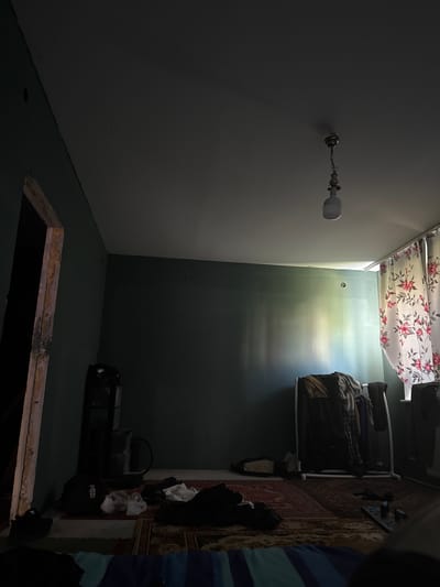 The image depicts an interior scene, likely a room in a dwelling. The perspective is skewed, making the orientation of the room appear disorienting. A ceiling light fixture with a white bulb hangs from the ceiling on the left side of the image, casting a dim light. To the right, a section of wall is painted a dark green color, and a white shelf unit is mounted on it. This shelf holds several folded garments, appearing to be clothing. Below the shelf, a red patterned rug is visible on the floor.

Further to the right, a person's arm and shoulder are visible, wearing a blue and white striped garment. On the floor in the lower right, a black bag with white lettering that reads "reebok" is placed. There is also a dark opening in the floor, possibly a trapdoor or a hole leading to a lower level, with wooden trim around its edges. The lighting suggests it is either nighttime or the room is poorly lit, with light primarily emanating from the single bulb and a faint gradient of light on the wall behind the shelf, hinting at a window or light source outside the frame. The overall impression is one of a cluttered and dimly lit living space. No specific activity is evident, but the presence of clothing and the general state of the room suggest it is a lived-in area.