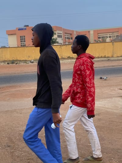 Two young men are walking along a roadside. One wears a red patterned hoodie and white pants with sandals. The other is in a black hoodie and blue pants, holding a white object. A building is in the background, and the sky is light. The ground appears dry and dusty. A road runs between the two figures and the background. The image is rotated 90 degrees counterclockwise.