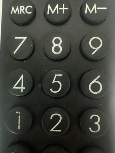 A close-up photographic image depicts a portion of a black electronic keypad, observed in the city of Pamidovo, Bulgaria. The keypad features circular black buttons with white alphanumeric characters. Visible buttons include "MRC", "M+", and "M-" in the uppermost row, followed by numerical buttons arranged as "7", "8", "9", "4", "5", "6", and "1", "2", "3" in subsequent rows. The white lettering exhibits minor wear and some specks of light-colored dust are present on the buttons and surrounding surface. No individuals or active events are depicted within the frame.