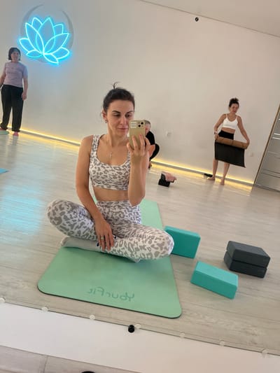 In a bright, modern yoga studio with light-colored wooden floors and white walls, a woman in a leopard-print yoga set is taking a selfie while sitting cross-legged on a mint-green yoga mat. She holds a smartphone in her right hand. To her left, another woman in a lavender t-shirt and black pants stands near a bright blue neon lotus flower sign on the wall. In the background, a woman in a white sports bra and black leggings is unrolling a yoga mat. Several yoga blocks are placed on the floor. The scene is reflected in a large mirror that covers the lower portion of the wall. The location of Aksu, Kazakhstan is not indicated in the image.