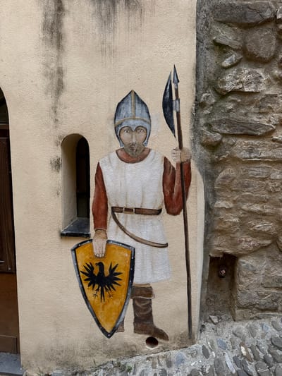 A mural of a medieval warrior stands on the stucco wall of a building in Dolceacqua, Italy. The warrior wears a helmet, a white tunic over a reddish-brown long-sleeved shirt, a brown belt, and brown boots. He holds a shield emblazoned with a black eagle on a yellow background in his left hand and a spear in his right. A small arched window is visible to the warrior's left. The wall to the warrior's right is constructed of rough stone. The ground in front of the wall is paved with cobblestones. The lighting suggests it is daytime.