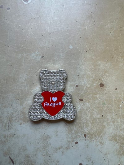 In Neratovice, Czechia, a silver-colored, bear-shaped decorative item, adorned with numerous small, clear, faceted rhinestones, is positioned on a light beige, distressed surface. The bear holds a central red heart emblem on its chest, featuring the white inscription "I ❤️ Prague". The background surface exhibits multiple fine scratches and scattered dark specks, indicative of wear. No individuals or other objects are visible within the frame.