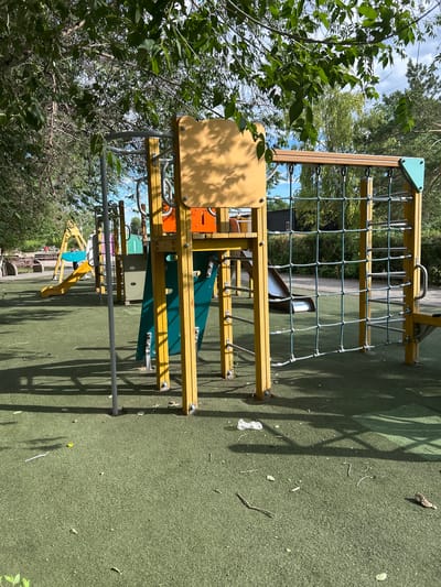 This is a medium-angle, outdoor photograph of a children's playground. The playground equipment is primarily yellow and green, with a climbing net made of thick rope. Various slides and platforms are visible, along with metal bars and supports. The ground surface is a textured, green material, likely a safety surfacing. The scene is bathed in sunlight, casting shadows from the playground structure and nearby trees onto the ground. Lush green foliage from trees overhangs the playground from the left side of the frame, suggesting a natural, wooded environment. The sky is visible in the upper left, with scattered white clouds. The overall impression is a bright, sunny day in a recreational outdoor space. There is no text visible in the image.