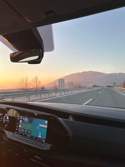 This image presents a first-person view from inside a vehicle traveling on a multi-lane highway at sunset, located near Thermopigi, Serres regional unit, Greece.

The vehicle's dark-colored dashboard occupies the lower portion of the frame. A central infotainment screen displays a navigation map indicating a route to "Serres - Synoriakou S", with a current distance of "8.4 mi" on a "Motorway". The screen shows "16:44" as the current time and "19:10" for estimated arrival. A digital instrument cluster to the left of the infotainment screen displays "17:39". The climate control system indicates a temperature setting of "23.0" degrees Celsius. A rearview mirror is centrally positioned at the top of the windshield.

Outside the vehicle, a paved highway with at least two lanes extends into the distance. A metal guardrail runs along the left side of the highway, behind which a sparse line of trees with bare branches is visible. Further along the left shoulder, a large, rectangular road sign structure, currently blank, is erected. Distant mountains, appearing hazy and multi-layered, form the horizon. The sky exhibits a distinct sunset gradient, transitioning from bright orange and yellow hues near the horizon to a lighter blue overhead. A green road sign is faintly visible in the far distance on the right side of the highway. No human subjects are discernible within the frame.