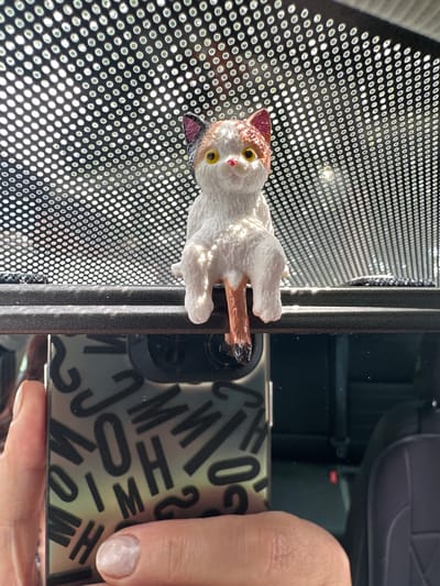 An ornamental figurine of a calico cat is perched on a dark gray trim, possibly the edge of a car's interior. The cat is white with patches of brown and black on its head and tail, and has yellow eyes and a pink nose. Behind the cat, a perforated dark surface is visible, with sunlight filtering through the perforations, creating a pattern of light and shadow. In the foreground, a hand is holding a smartphone, which is reflecting an interior car scene. The smartphone case is black and features a repeating pattern of letters, including "J", "M", "O", and "H". The background of the smartphone reflection shows dark seats and possibly a seatbelt. The scene appears to be inside a vehicle. The location is Prien am Chiemsee, Germany.