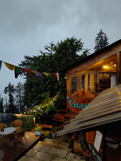 The image depicts an outdoor seating area and the exterior of a wooden structure, likely a cafe or guesthouse, in Siumsa, India, under an overcast sky. Prayer flags in various colors, including yellow, green, red, blue, and white, are strung across the scene. String lights are visible, illuminating trees and the building. The wooden structure has large windows, some with yellow curtains, and a sign with teal-colored lettering that appears to read "GLAM." Inside the building, bookshelves are visible through the windows.
In the foreground, there is a wet wooden deck with a small round table and two wicker chairs. Adjacent to the deck is a stone patio, also wet, suggesting recent rainfall. Several potted plants are scattered around the area, including a small green plant in a grey pot and a red flowering plant in a brown pot. The ground is visibly wet and reflective. The overall atmosphere is muted, possibly due to the cloudy weather. No people are visible in the image. The location appears to be a scenic spot, possibly in a mountainous or hilly region given the presence of tall evergreen trees in the background.