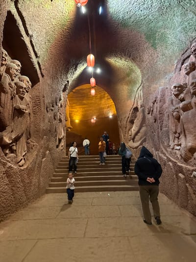 Captured in Shizhong, China, the scene portrays a stone tunnel adorned with carved figures on its walls and ceiling. A series of red lanterns are suspended along the tunnel's length. A group of people is ascending a stone staircase. One person, dressed in a black jacket and olive pants, stands towards the top of the staircase.
