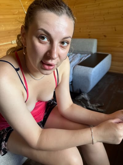 A young woman with blonde hair tied back is shown from the chest up, looking towards the camera. She is wearing a pink tank top with dark straps and a patterned skirt. She has on earrings and a necklace. Her arms are resting on her lap. In the background, a sofa with a dark top is visible, and the room appears to be made of wood. The scene is indoors, likely in a residential setting.