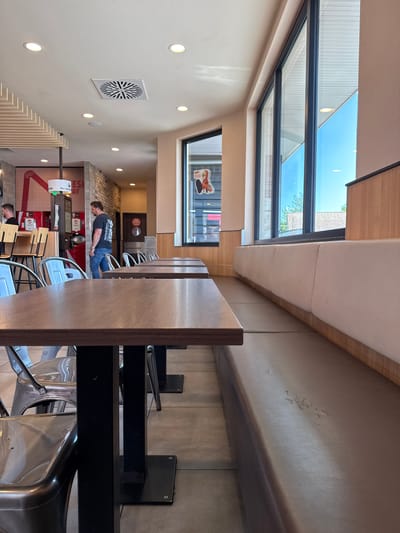 The image depicts the interior of a fast-food restaurant in Mülheim-Kärlich, Germany, with a focus on the seating area. In the foreground, several empty, dark brown wooden tables are arranged in a row, with one table closest to the camera being the most prominent. These tables have black metal legs secured to the tiled floor. Beside the tables, there are metallic chairs, some of which are also empty.

To the right of the tables, a long, curved bench seat upholstered in a brown, faux-leather material is visible. The bench shows signs of wear, with some peeling and discoloration on its surface. This seating arrangement runs along a large window that offers a view of the bright blue sky and some trees and buildings outside. The window is framed by dark grey metal.

Further into the restaurant, a man in a black t-shirt and blue jeans is walking away from the camera, heading towards a counter area. Behind him, there are additional tables and chairs. In the background, a counter with stools is visible, and a red graphic design with the text "ES AY" is present on a wall. Above, a ventilation grate and circular lights are integrated into the white ceiling. The overall environment suggests a typical, modern fast-food establishment.