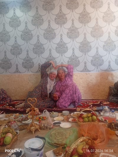 Two women are seated on a patterned rug in front of a wall with a repeating damask-style wallpaper pattern. The woman on the left is wearing a dark floral dress and a light-colored headscarf, while the woman on the right is wearing a purple floral dress and a patterned headscarf. Both women are forming heart shapes with their hands above their heads. In front of them is a low table covered with a rug, laden with food and drinks. Visible items include fruit baskets containing apples, bananas, and grapes, platters of flatbread, bowls of nuts and candies, a teapot, a bottle of soda, and various other dishes. A gold-colored tiered stand with a heart at the top is also present on the table. The overall scene appears to be a gathering or meal. The image also contains a watermark in the bottom left corner that reads "POCO C75" and a date and time stamp in the bottom right corner: "16.07.2026 14:34". The location of Чоң-Гара, Kyrgyzstan is mentioned in the prompt.