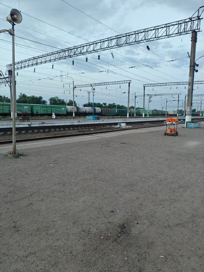 This image captures a view of a train station, likely in the city of Shu, Kazakhstan, characterized by its overcast sky and extensive railway infrastructure. The foreground is dominated by a gravelly platform, with a subtle patch of worn-down ground in the lower center. To the left, a tall pole supports a large loudspeaker, a common fixture at such locations. Beyond the platform, a set of train tracks runs parallel, with a train of green freight cars and white tank cars visible in the middle ground, parked on the tracks. Overhead, a complex network of electrical wires and support structures crisscrosses the sky, indicating the presence of an electrified railway system. On the platform in the middle ground, a small, two-wheeled cart is visible, laden with numerous orange and yellow bags, suggesting the presence of vendors or goods being transported. The overall atmosphere is one of a functional, somewhat utilitarian transportation hub under a muted sky.