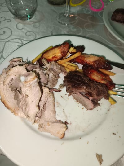 The image shows a white dinner plate with various types of meat, french fries, and some remnants of sauce on a table with a patterned tablecloth. On the left side of the plate, there are large slices of roasted meat, possibly pork. In the center and right, there are pieces of roasted chicken or duck, glazed with a dark sauce, and interspersed with french fries. A piece of dark roasted beef is also visible on the right side of the plate, with a fork resting on it. In the background, out of focus, are wine glasses and colorful plastic rings, suggesting a festive or celebratory gathering. The scene appears to be indoors, likely in a restaurant or a home dining area, set in the city of Vinchiaturo, Italy.