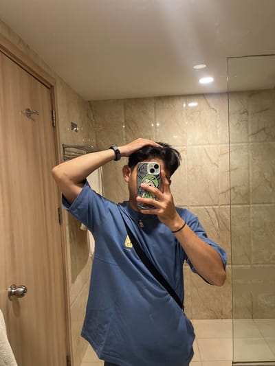 A young man stands in a bathroom, taking a selfie in a mirror. He is wearing a blue t-shirt and has his hair tousled with one hand. A black watch is visible on his left wrist, and a black string bracelet is on his right wrist. A necklace with a pendant is also visible. His phone, with a cartoon sticker on its case, is held up to take the picture. The bathroom features beige tiled walls, a wooden door with a silver handle, and a glass shower enclosure. The scene appears to be inside a hotel or residential building. The location is the city of Dumai, Indonesia.