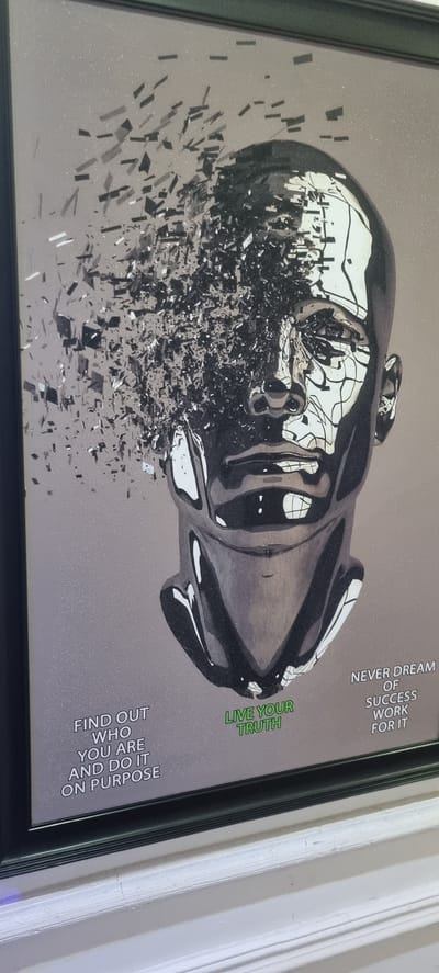 A framed motivational poster is displayed at an angle, likely hanging on an indoor wall, with a segment of a white wall visible on the right side. The central artwork features a highly reflective, dark metallic sculpture or rendering of a human head in profile, facing towards the right. The upper portion of the head, from the forehead upwards, appears to be disintegrating into a multitude of small, dark, angular fragments that scatter downwards and to the left across the light grey, subtly glittery background. The lower part of the face, including the nose, mouth, chin, and jawline, remains intact and exhibits a polished, mirror-like finish.

The poster includes three distinct motivational texts. In the upper right corner, oriented vertically, is the white text: "NEVER DREAM OF SUCCESS WORK FOR IT". Below this, centrally located on the right, the phrase "LIVE YOUR TRUTH" is presented in green text. In the lower left corner, also vertically oriented, the white text reads: "FIND OUT WHO YOU ARE AND DO IT ON PURPOSE". The poster is enclosed within a simple black frame. The location context provided for this image is Aiyetoto-Asogun, Nigeria.