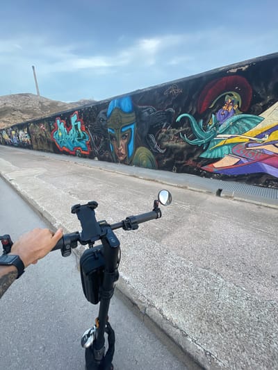 The image shows a first-person perspective of someone riding an electric scooter along a paved path. On the right side of the path is a long, graffiti-covered wall, featuring colorful murals of various figures, including a warrior with a blue helmet, a wolf, and an elephant-like creature. The background reveals a slightly hilly landscape with a tall, industrial chimney in the distance, characteristic of the industrial port area of Cartagena, Spain. The sky is overcast with light clouds. The scooter handlebars are visible in the foreground, with a phone mount, a rearview mirror, and a small bag attached. A tattooed arm and hand are gripping the right handlebar. The ground is a rough, grey asphalt with concrete curbs.