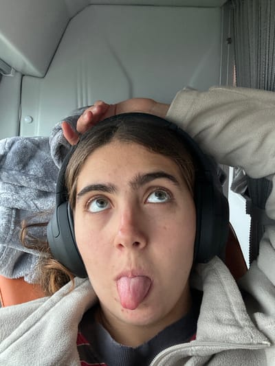 A woman is seen from a low angle, tilting her head back to face the camera. She is wearing headphones and a gray, fluffy jacket. Her eyes are directed upwards and to the left, while she sticks her tongue out of the side of her mouth. The background is a gray, seemingly interior space, possibly a vehicle or room with light gray walls and furniture. The overall impression is casual and playful.