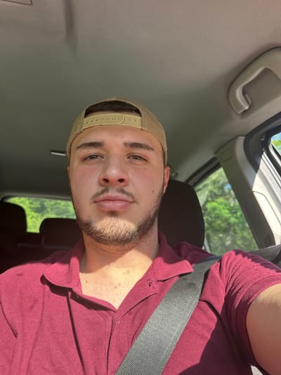 The media file is a portrait of a young man sitting in the passenger seat of a car. He is wearing a maroon polo shirt and a tan baseball cap. His face is turned towards the camera, and he has a slight smile. The man has dark hair and brown eyes, and he is clean-shaven except for some stubble on his chin and upper lip. The car's interior is visible in the background, including the headliner, a grab handle, and a seatbelt. Sunlight is streaming in from the open sunroof, illuminating the man's face and casting shadows. Outside the car, green trees and blue sky are visible, suggesting it is a sunny day. The overall mood of the image is casual and relaxed.
