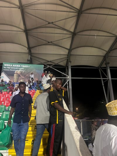 The image depicts a nighttime scene within the interior stands of a stadium or arena in Zanzibar City, Tanzania. Artificial lighting illuminates the area, revealing a crowd predominantly composed of men.

In the foreground, a man in a dark blue short-sleeved collared shirt and blue jeans descends yellow-colored stairs on the left, holding a green bag in his left hand. To his right, a man in a grey t-shirt and blue jeans, wearing a white and blue patterned kufi, ascends the same stairs, with his back partially turned. Further to the right, a man wearing a black and yellow striped short-sleeved shirt with "ZAN" printed on it and black track pants with red stripes leans against a concrete railing, looking left. A partially visible man in a white garment and a white and yellow patterned kufi stands on the far right, facing away from the viewer.

The midground features multiple rows of stadium seating, with both red and green plastic seats occupied by numerous individuals, many of whom wear kufis or other head coverings. A large green banner is visible on the upper left wall, bearing text in Swahili: "WEKA AKIBA YAKO!" (Deposit your savings!) and "Na PBZ IKHLAS haina makato Mwisho wa Mwezi" (And PBZ IKHLAS has no end-of-month fees). The banner includes an illustration of a hand dropping coins into a house and a man speaking into a microphone. Above the seating area, a light-colored, arched fabric roof is supported by a grey metal truss structure. Yellow stairs with "VIP" markings and numbering (e.g., "91") are present throughout the stands. The background beyond the stadium structure is dark, indicating the nighttime setting.