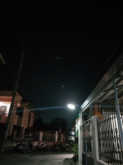 The image depicts a street scene at night in San Pascual, Philippines. The sky is dark, with a streetlamp in the foreground illuminating a section of a paved area. To the left, a multi-story building with a balcony is visible, adjacent to a utility pole with overhead wires. In front of the building, a three-wheeled vehicle, resembling a tricycle or an auto-rickshaw, is parked, alongside two motorcycles and another motorcycle further down the street.
The right side of the image features a covered patio area with a corrugated metal roof supported by metal posts. Clothes are hanging to dry on a clothesline beneath the roof. A black metal gate with decorative elements stands in front of a white pillar. Beyond the gate, parts of a house are visible. In the background, more houses, fences, and some greenery can be seen, along with the faint outline of trees. A distant vehicle's headlights are visible in the far background.
In the dark night sky, several faint lights are visible. A bright circular object, likely the moon or a planet, is visible slightly above and to the left of the streetlamp. A horizontal line of small green lights is also present in the upper portion of the sky, possibly distant lights or an aerial phenomenon. The overall atmosphere is quiet and residential, suggesting a typical evening in a Philippine town. There is no visible text in the image.