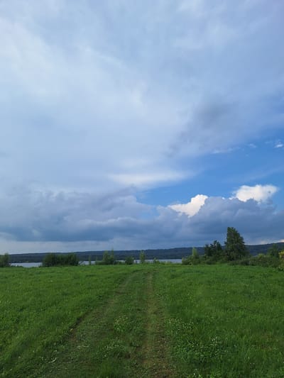 The image depicts a vast expanse of green grass with a faint dirt path winding through it, leading towards a body of water in the distance. The sky above is a dramatic mix of bright blue and heavy, dark clouds, suggesting an impending storm or recent rainfall. A line of trees and hills forms the horizon behind the water. The scene appears to be a natural landscape, likely in a rural or park-like setting. Based on the provided information, this location is identified as Atamanovka, Russia. There are no people or animals visible in the image.