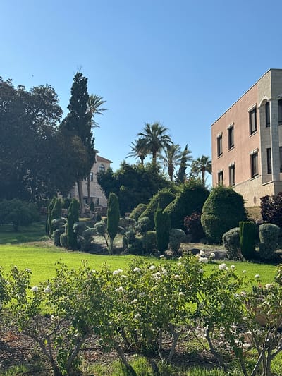 The image depicts a well-maintained garden with a building in the background, located in Emek HaYarden Regional Council, Israel. The foreground is dominated by a vibrant green lawn, bordered by a rose bush with white flowers. Behind the lawn, a meticulously landscaped garden features an array of topiaries, cypress trees, and various shrubs. In the mid-ground, a pale pink building with stone accents stands out, featuring multiple windows. Further in the background, another building constructed of stone is partially visible, surrounded by lush trees, including palm trees, characteristic of the region. The sky is a clear, bright blue. There are no people visible in the scene.