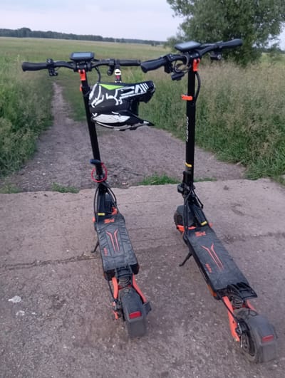 Two electric scooters, likely Kukirin G2 Pro models given the visible branding on the handlebars, are parked on a dirt and concrete path. The scooters are black with orange accents. One scooter has a helmet with a white and black graphic design hanging from its handlebars. Both scooters are facing away from the viewer. The path is flanked by tall grass and a field, with a line of trees in the background. The sky is overcast. The scene appears to be in Wiktorów, Poland, based on the provided context.