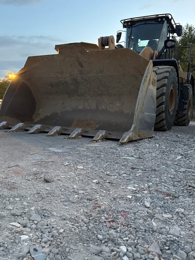 A low-angle, close-up shot focuses on the front of a large, yellow Caterpillar wheel loader parked on a gravel surface. The loader's massive bucket, partially filled with dirt, dominates the foreground. The bucket's edge has several prominent teeth, some of which appear worn and embedded with debris. Behind the bucket, the loader's large, heavy-duty tires are visible, showing a rugged tread pattern. The cab of the loader, with its tinted windows, is visible in the upper right of the frame. Two yellow warning lights are illuminated on the top of the cab. In the background, a line of trees with green foliage is present. The sky above is a mix of pale blue and soft grey clouds, with a bright, warm light originating from the left side of the frame, likely from a streetlamp or the setting sun, casting a glow on the loader's bucket. The ground is covered in a mixture of small and medium-sized grey gravel, with occasional larger stones and fragments of what appear to be broken concrete or building materials scattered throughout. The scene suggests an outdoor industrial or construction setting, possibly at dusk given the lighting conditions.