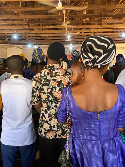 An indoor scene in Nev, Nigeria, depicts a gathering of people observed from behind. In the immediate foreground, from left to right, are three individuals: a person in a white short-sleeved top and dark trousers; a person in a black short-sleeved shirt adorned with orange, yellow, and green floral patterns; and a woman wearing a textured deep purple short-sleeved dress with a zippered back featuring a metallic 'B' charm. This woman wears gold hoop earrings, a black and white striped headwrap, and has a black purse with a gold chain strap over her shoulder. She is carrying a baby on her back, partially visible and looking towards the right. The overhead environment features an exposed wooden rafter ceiling, illuminated by multiple bare incandescent light bulbs, and includes two white ceiling fans. Numerous additional individuals, many with head coverings, are present in the blurred background.