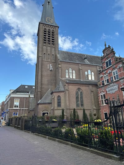 The image shows the Westerkerk church in Enkhuizen, Netherlands. The brick church has a tall clock tower and a sign reading "1901" on its side. The sky above is blue with scattered clouds. Adjacent to the church is a red brick building with white-framed windows. A black iron fence runs along the edge of the church property, bordering a brick-paved sidewalk. Beyond the fence, a row of colorful tulips and other greenery can be seen. In the foreground, a section of the brick sidewalk is visible, leading towards buildings with storefronts in the distance.