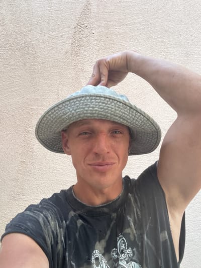 In Pelinovo, Montenegro, a man with light skin and short blonde hair is posing for a selfie outdoors against a textured, light-colored wall. He is wearing a light green bucket hat, pulled down to partially cover his forehead, and a dark t-shirt with a faded, abstract pattern. His right hand is raised, holding the brim of the hat. The man is smiling slightly, looking directly at the camera. The lighting suggests it is daytime, likely sunny, casting subtle shadows.