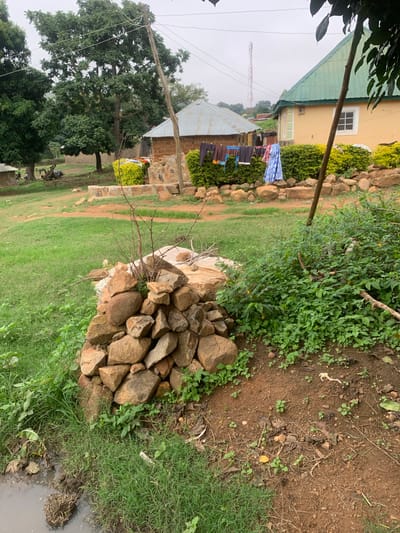 The image depicts a rural scene in Zhwarki Mile 10, Nigeria. In the foreground, a pile of rocks sits on a patch of grass near a muddy puddle. Behind the rocks, a dense green bush with small yellow flowers is visible. Further back, a cluster of traditional brick houses with corrugated metal roofs are arranged around a larger, yellow-painted house with a green roof. Clothes are hanging on a line between two of the houses. In the background, a tall communication tower is visible against a cloudy sky, with lush green trees surrounding the settlement. The overall impression is of a simple, functional living environment in a natural, semi-urban setting.