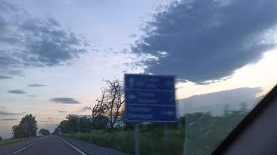 The image depicts a view from a vehicle traveling on a road. There is a road sign with the names of cities and distances. The environment includes trees, shrubs, and an open sky with cloud formations. The location mentioned is in the city of Chernograd, Bulgaria.
