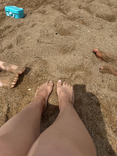This media file shows a close-up, top-down view of several pairs of feet partially submerged in sand on a beach. The sand is a mix of light brown and grey grains, with some small pebbles. In the foreground, a pair of bare feet with painted white toenails are visible, and further back, another pair of feet are also visible. Two other pairs of feet are also present, one of which is partially covered in sand. A small, bright turquoise toy, possibly a camera or a lock, is located in the lower left quadrant of the image. The lighting suggests it is daytime, and the shadows cast on the sand indicate the sun is at an angle. The scene appears to be a casual day at the beach, possibly a family outing. No visible text is present in the image.