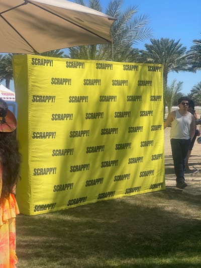 Here is a forensic description of the image:

**Scene:** The image depicts an outdoor event in Thermal, United States.

**Objects:**
*   A large, yellow backdrop printed with the word "SCRAPPY!" repeatedly in black.
*   An open-sided tent or awning, partially visible on the left side.
*   Palm trees in the background.
*   A patch of grass.
*   A small table with what appears to be merchandise or promotional items.

**People:**
*   Two individuals are visible standing behind the backdrop. One is wearing a white top and dark pants, with sunglasses.

**Environment:**
*   The sky is clear and blue. The light suggests it's daytime.
*   The presence of palm trees indicates a warm climate.