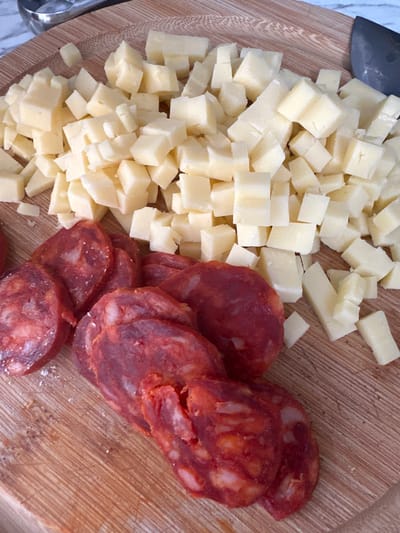 The image shows a close-up, overhead shot of diced cheese and sliced chorizo on a wooden cutting board. The cheese is light yellow and cut into small, irregular cubes, piled in the upper right portion of the frame. The chorizo slices are circular with a deep red color and visible white fat marbling, arranged in the lower left of the image. The wooden cutting board has a visible grain pattern and some scattered crumbs. In the upper left corner, a small portion of a metal utensil is visible. There are no people or indications of a city location.