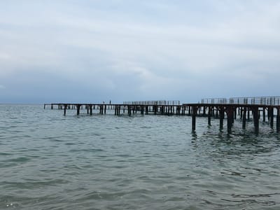 The image shows a long, weathered pier extending over calm, grey water under a cloudy sky. Several figures are visible on the pier, silhouetted against the sky. The pier is supported by numerous wooden pilings that plunge into the water. In the foreground, the water is slightly rippled, and a faint reflection of the pier is visible beneath the surface. The scene appears to be a public access point on a body of water, likely a lake or the sea, in or near Kara-Oy, Kyrgyzstan.