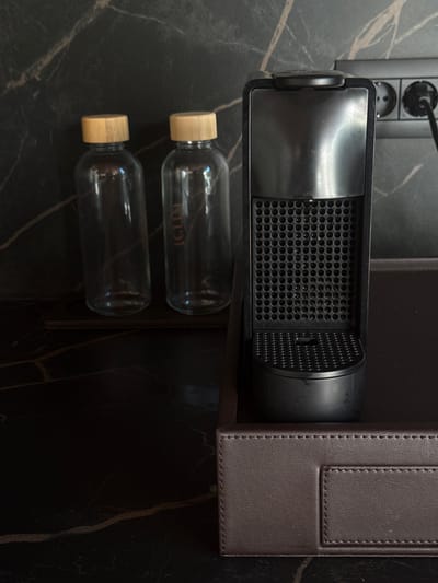 A close-up, eye-level shot captures a coffee machine and two glass bottles with wooden stoppers on a dark, marble-patterned surface. The coffee machine, a Nespresso model, is black and positioned on a brown leather tray. To its left, the two identical glass bottles are standing upright. The background features a dark, veined marble wall with electrical outlets visible on the right side, one of which has a black power cord plugged in. The scene appears to be in Bakovići, Montenegro.