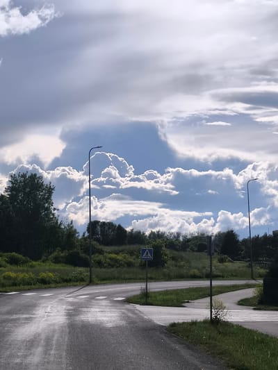 The image captures a dynamic sky over a semi-urban landscape in Tänassilma, Estonia. Towering cumulonimbus clouds, illuminated by the sun, dominate the upper portion of the frame, their edges glowing with bright white light against a deep blue sky. The lower part of the image shows a paved road, wet and reflective, suggesting recent rain. A pedestrian crossing is clearly marked on the road. To the left of the road, a lamppost stands tall, its dark silhouette contrasting with the bright sky. A small blue and white pedestrian crossing sign is visible on a post in the middle ground, along with other traffic signs. Green grass and trees populate the landscape, forming a verdant backdrop to the road and the dramatic sky. The scene appears to be a quiet intersection on the outskirts of the town.